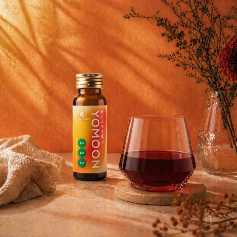 Brown bottle with a gold cap and orange label beside a glass of red beverage on a warm orange background, still-life setup.