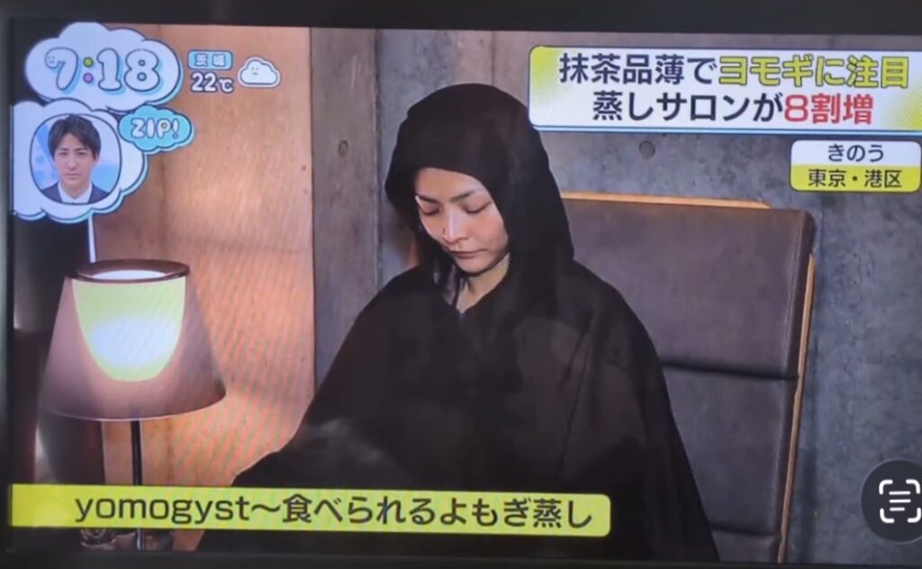On a Japanese TV show, a woman wearing a black hooded cloak sits in a chair beside a lit lamp; a yellow caption at the bottom reads 'yomogyst〜食べられるよもぎ蒸し' while other on-screen graphics appear in the corners.